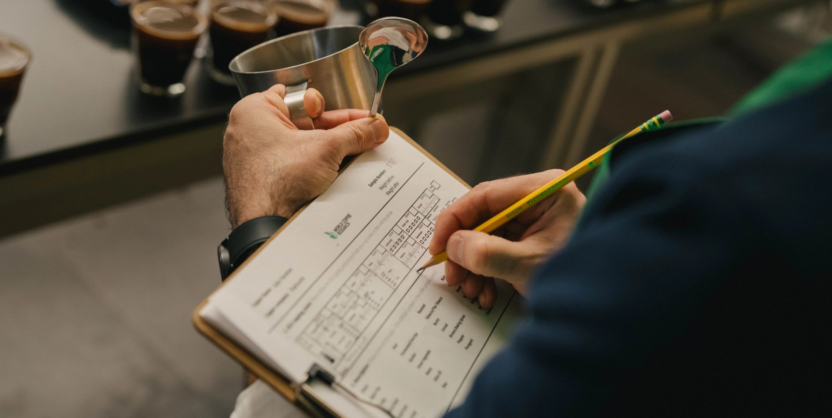 Person taking notes with a pen and a notebook, holding a metal cup, in a kitchen setting.