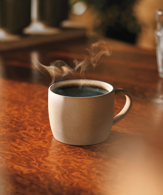 A steaming cup of black coffee in a stoneware mug on a polished wooden table.