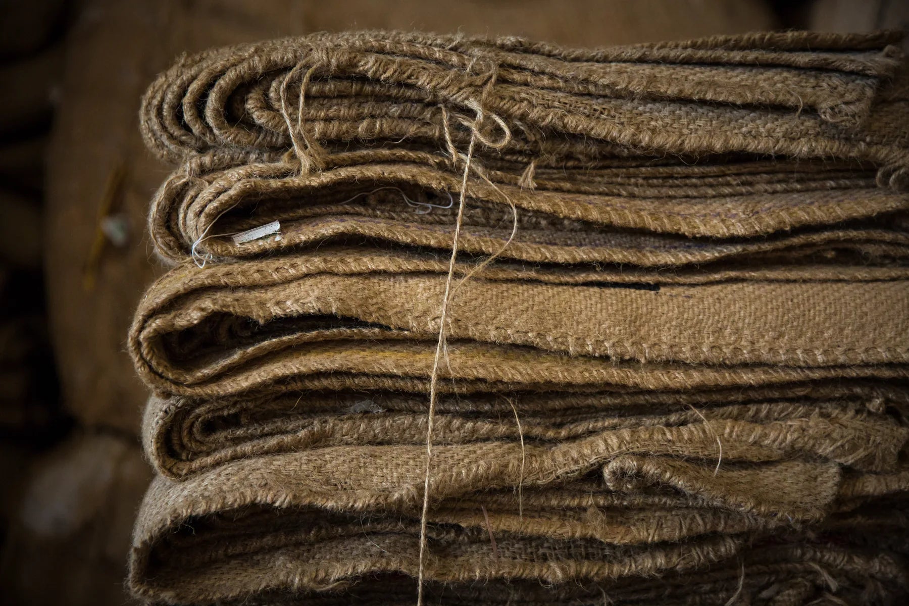 A close-up stack of folded natural brown jute or burlap fabric sheets tied together with a string, showing the coarse woven texture.