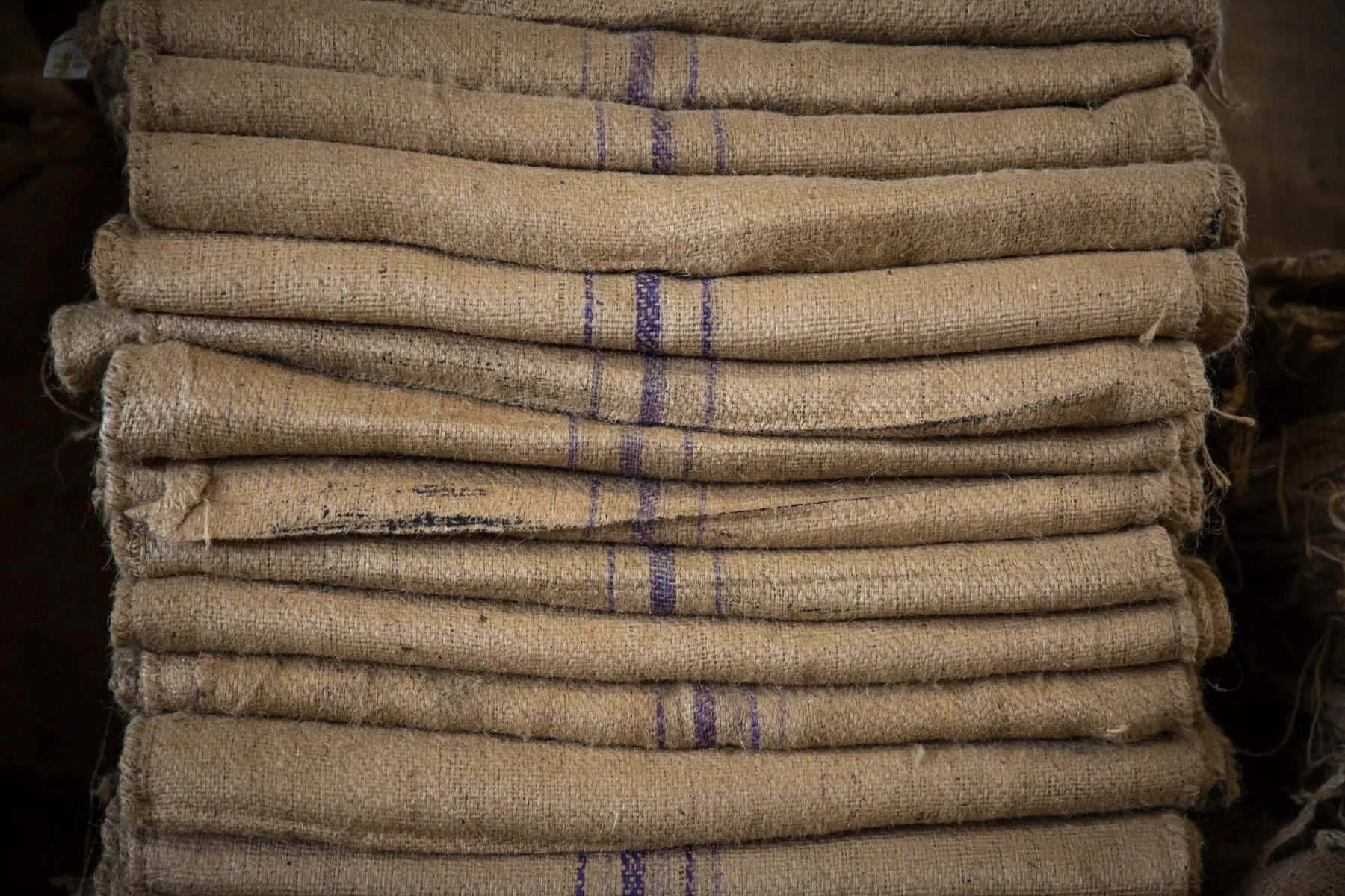 A close-up stack of natural brown burlap sacks with purple woven stripes, tightly folded and stacked together.