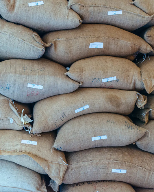 A close-up view of stacked natural burlap coffee sacks filled with goods, each marked with a small white paper label and handwritten numbers.