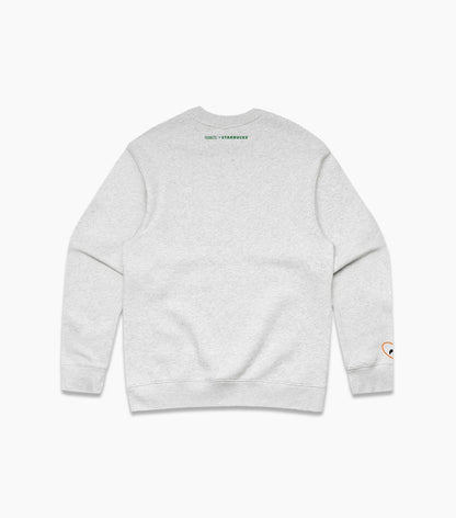 A light heather gray crewneck sweatshirt shown from the back, featuring a small green 'PEANTS + STARBUCKS' logo printed at the upper neckline and a small multicolor graphic on the left sleeve cuff.