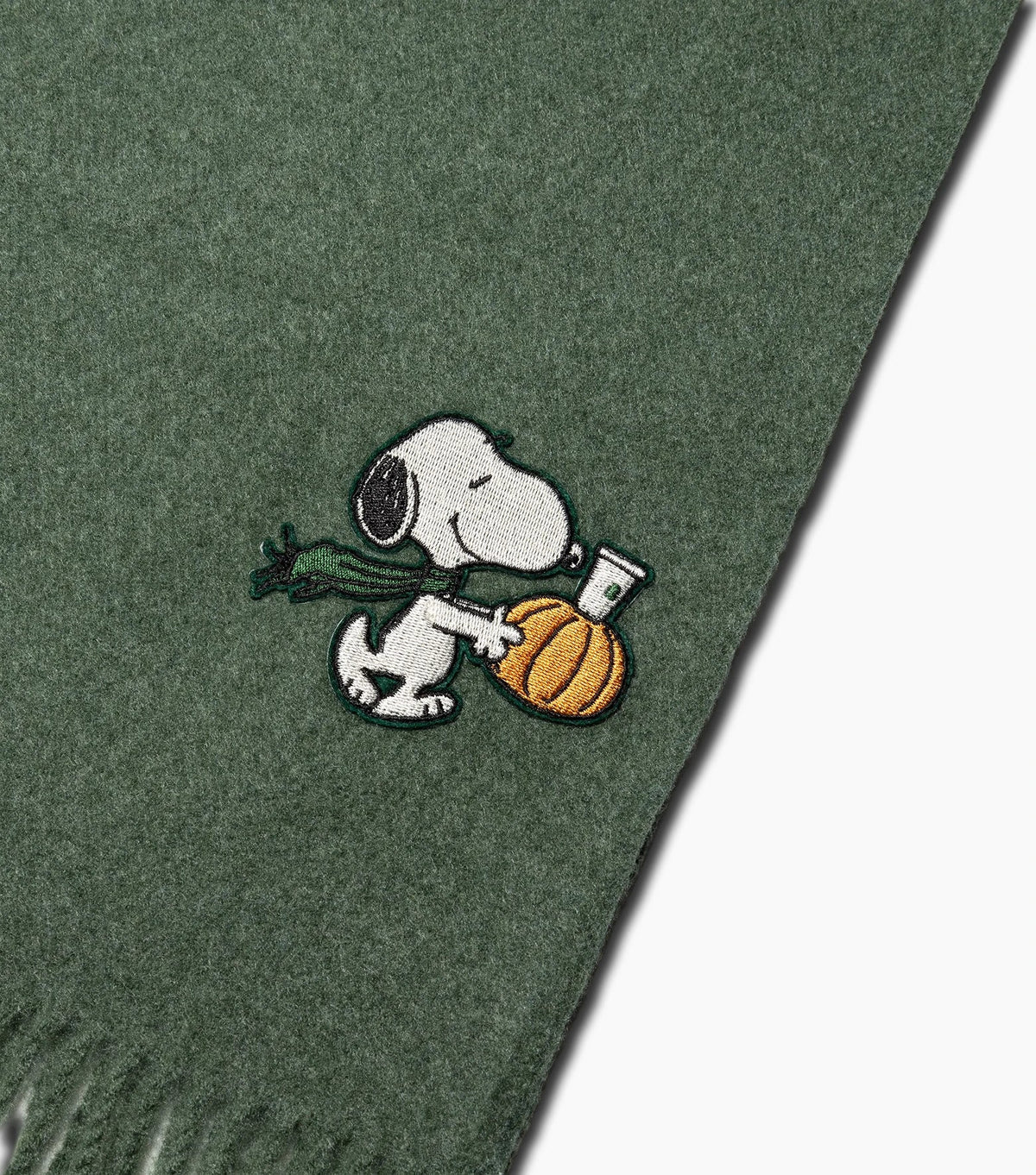 A close-up of a dark green knit scarf featuring an embroidered patch of Snoopy holding a green turnip and an orange pumpkin.