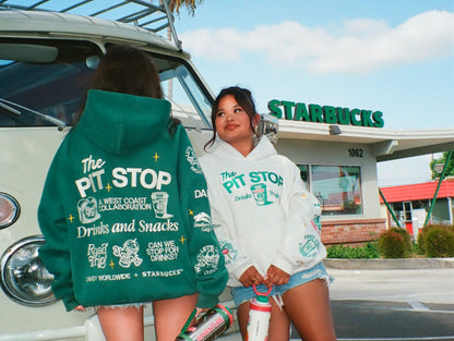 Two models wearing Dandy + Starbucks-branded hoodies stand in front of a white vintage van and a Starbucks building. One model shows the back of a dark green hoodie with white graphic text, while the other shows the front of a white hoodie with matching graphics.