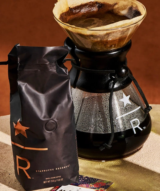 A bag of Starbucks Reserve whole bean coffee next to a glass pour-over brewer with the Starbucks Reserve logo.