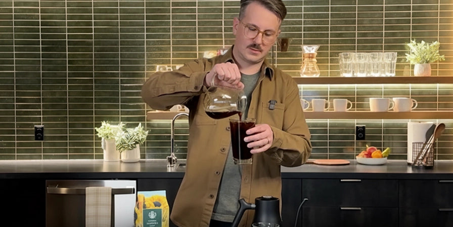 Man in a kitchen pouring a drink, with a tiled wall and kitchenware in the background.