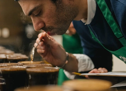 Person in a green apron tasting coffee from a spoon