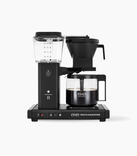 A black Moccamaster coffee maker with a clear water reservoir, a black filter basket, and a glass carafe containing brewed coffee, shown on a clean white background.