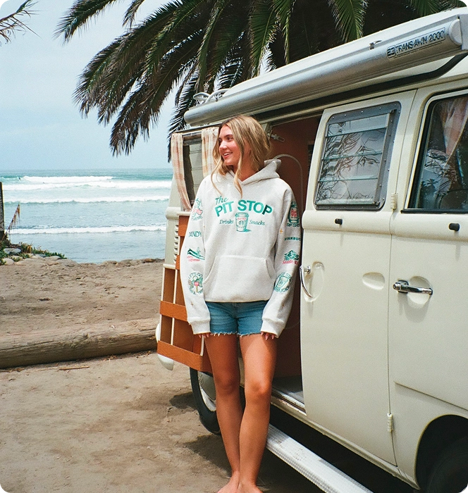 A woman stands barefoot on a beach next to a cream-colored vintage VW bus, wearing a white hoodie with teal 'PIT STOP' graphics and denim shorts.