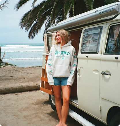 A woman stands barefoot on a beach next to a cream-colored vintage VW bus, wearing a white hoodie with teal 'PIT STOP' graphics and denim shorts.