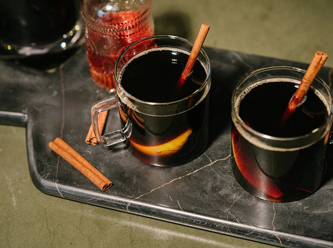 Two mugs of hot mulled wine with cinnamon sticks on a dark tray.