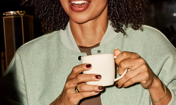 A close-up lifestyle shot of a person holding a white ceramic Starbucks mug, wearing a sage green cardigan and brown top.