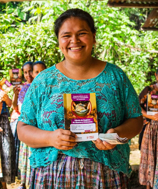 Norma smiles as she holds her savings and loans booklet, with other women from her group holding the same booklet behind her.