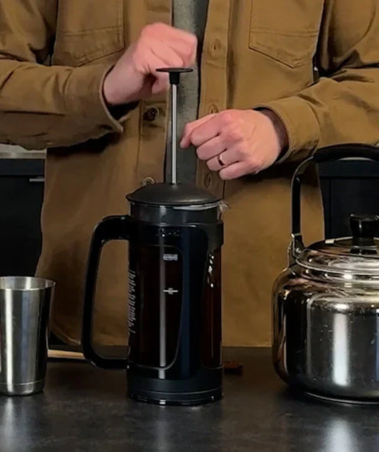 Person standing behind a range of coffee making tools on the table, with a coffee press in the middle, a metal cup to its left and a kettle on the right.