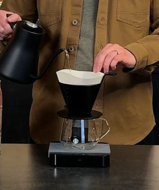 Person pouring hot water from a kettle, through filter paper into plastic pour over on top of a glass mug.