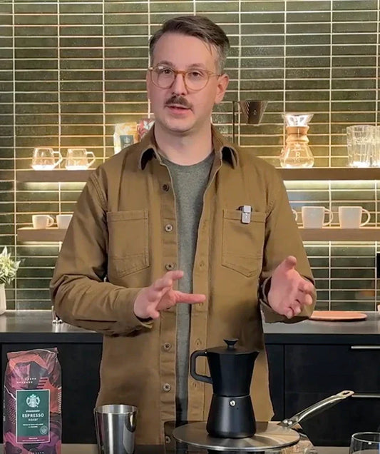 The barista, Lincoln in a kitchen setting, with a moka pot in front of him, alongside with a bag of Starbucks Espresso Coffee and a metal cup next to it.