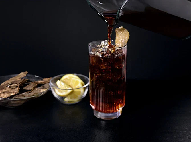 Coffee being poured into a glass of ice garnished with candied ginger, with dried lemon peel and lemon in the background.