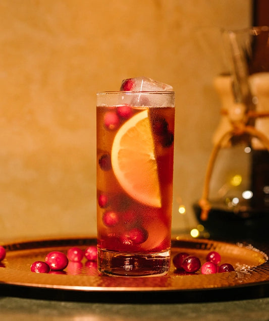 A tall glass of an iced drink with an orange slice and cranberries, on a copper tray with more cranberries around it.