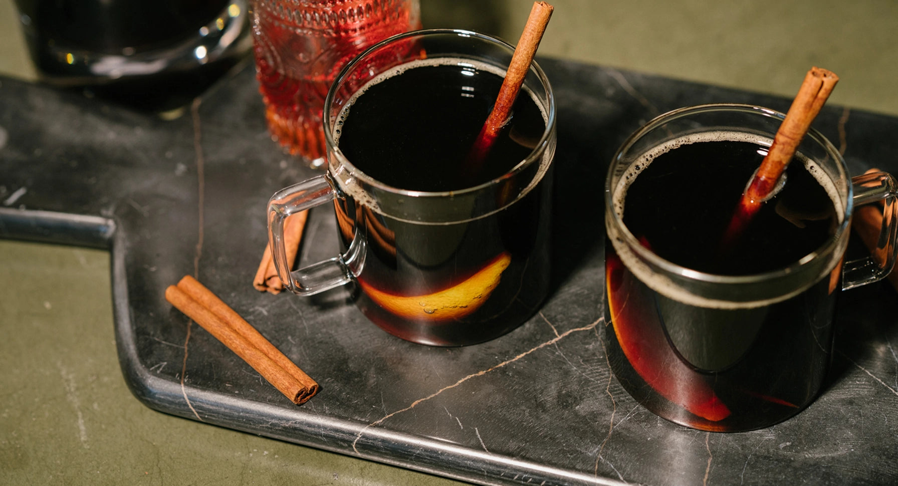 Two mugs of mulled wine with cinnamon sticks on a dark tray.