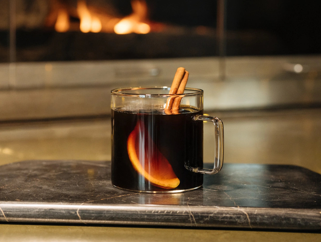 Glass mug of mulled wine with cinnamon sticks on a stone surface in front of a fireplace.