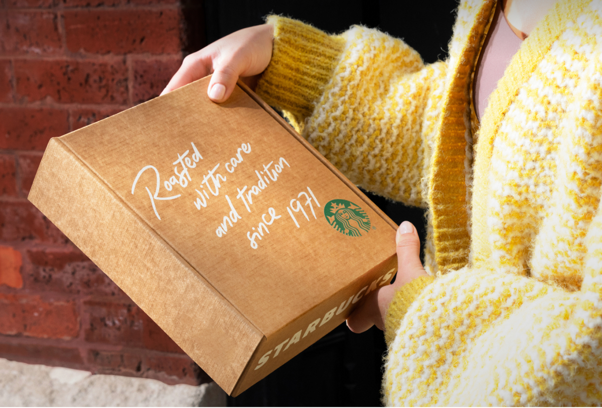 A person in a yellow knit cardigan holds a brown cardboard Starbucks box outdoors against a red brick wall. The box has white script text reading 'Roasted with care and tradition since 1971' and the green Starbucks siren logo on the top, with 'STARBUCKS' visible on the side.